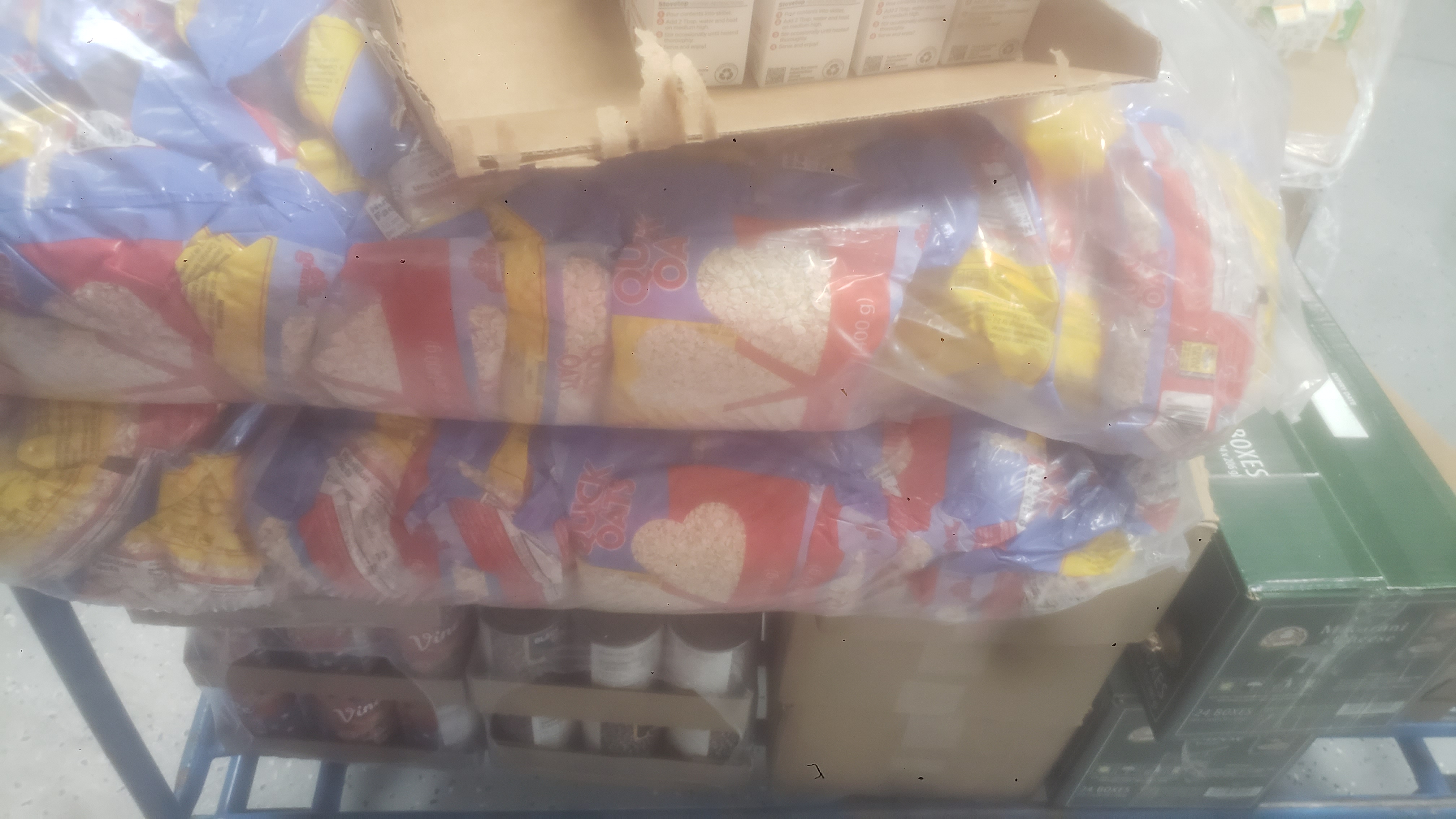 Box of shelf-stable meals on top of bags filled with oats. Under that is cans of tomato sauce, applesauce, and black turtle black beans which is next to boxes of mac n cheese on a shelf cart.
